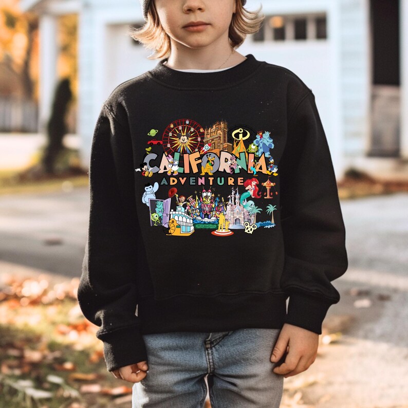 May include: Black sweatshirt with the words "CALIFORNIA ADVENTURE" in colorful letters and various cartoon illustrations. The design includes a Ferris wheel, castle, and other theme park imagery. The sweatshirt is worn by a person.