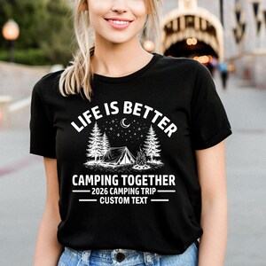 Puede incluir: Camiseta negra con el texto blanco "LIFE IS BETTER CAMPING TOGETHER". El dise&ntilde;o incluye una tienda de campa&ntilde;a, &aacute;rboles, una fogata, una luna creciente y estrellas. Debajo est&aacute; escrito "2026 CAMPING TRIP - CUSTOM TEXT".
