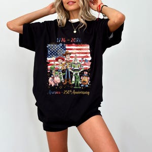 Op de afbeelding: Zwart t-shirt met een afbeelding van de Amerikaanse vlag, stripfiguren en de tekst "1776-2026 America - 250th Anniversary". Het ontwerp bevat vuurwerk en sterren.