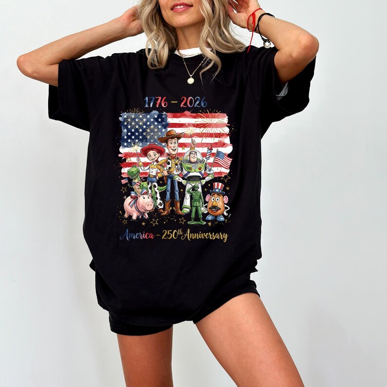 Op de afbeelding: Zwart t-shirt met een afbeelding van de Amerikaanse vlag, stripfiguren en de tekst "1776-2026 America - 250th Anniversary". Het ontwerp bevat vuurwerk en sterren.