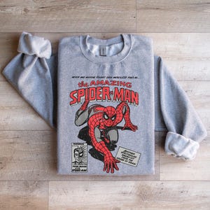 May include: A gray sweatshirt with a vintage comic book design featuring Spider-Man. The text reads "NEVER HAS ANYONE FOUGHT SUCH MERCILESS FOES AS THE AMAZING SPIDER-MAN".