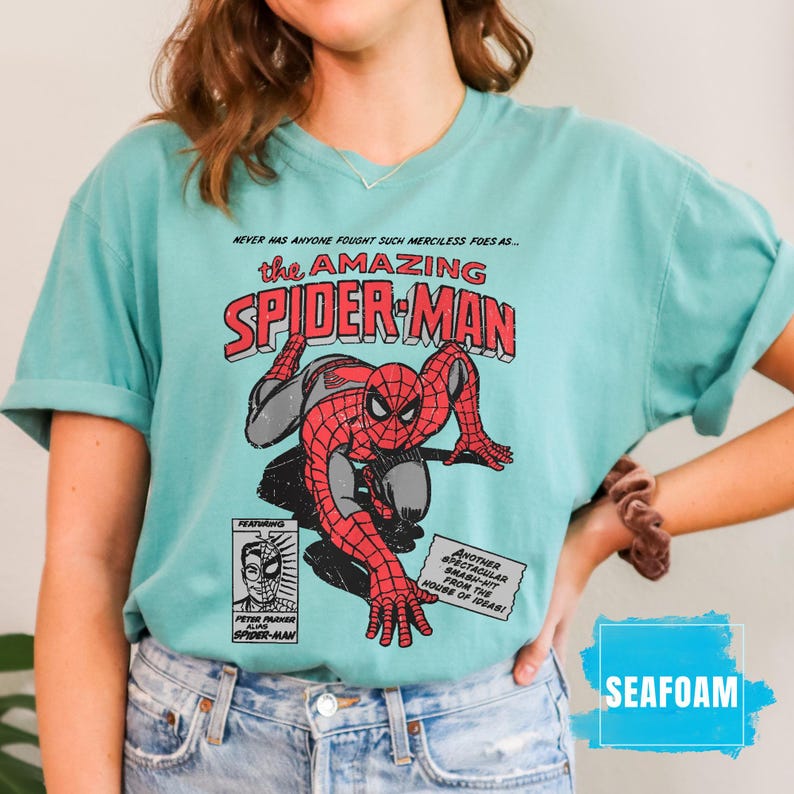 May include: Seafoam green t-shirt featuring a vintage comic book graphic of Spider-Man. The design includes the text "the AMAZING SPIDER-MAN" and other comic book elements. The shirt is a casual style.