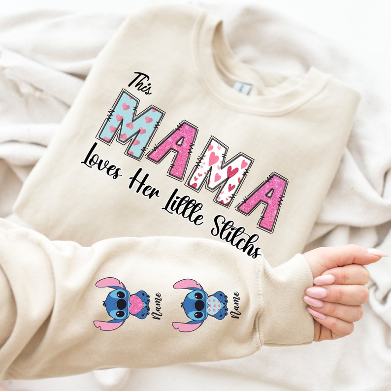 May include: A beige sweatshirt with the text "This Mama Loves Her Little Stitchs" in pink and blue letters with hearts. The sleeves have a blue Stitch character with pink hearts.