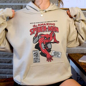 May include: A beige hoodie with a vintage comic book design featuring Spider-Man in a red and blue suit. The text on the comic book cover reads "The Amazing Spider-Man".