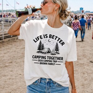 Puede incluir: Camiseta blanca de manga corta con el texto negro "LIFE IS BETTER CAMPING TOGETHER". El dise&ntilde;o incluye una tienda de campa&ntilde;a, &aacute;rboles y una fogata. Debajo est&aacute; escrito "2026 CAMPING TRIP - BROWN FAMILY".