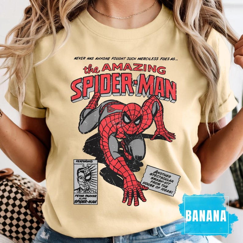 May include: A light yellow t-shirt featuring a comic book style illustration of Spider-Man in red and gray. The text "the AMAZING SPIDER-MAN" is above the image. Additional text includes "NEVER HAS ANYONE FOUGHT SUCH MERCILESS FOES AS..." and "FEATURING PETER PARKER ALIAS SPIDER-MAN".