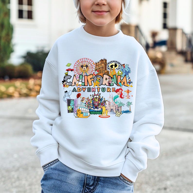 May include: White sweatshirt with a colorful graphic that says "CALIFORNIA ADVENTURE". The graphic includes cartoon images of a Ferris wheel, Mickey Mouse, a castle, and other amusement park elements. The sweatshirt is worn by a person.