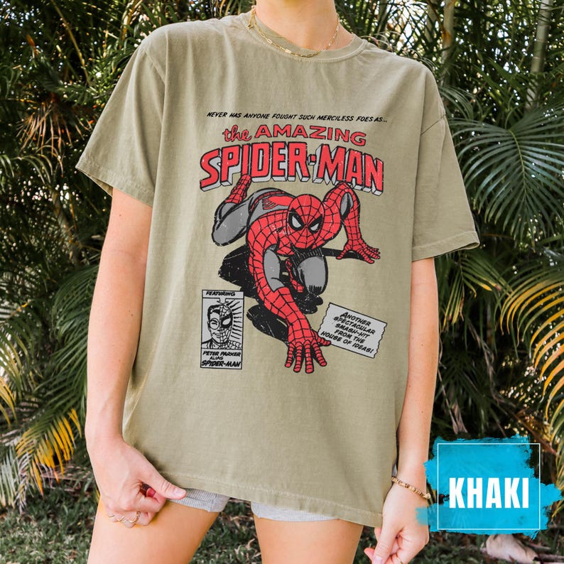 May include: Khaki-colored t-shirt featuring a comic book graphic of Spider-Man. The design includes the text "the AMAZING SPIDER-MAN" in red and white, with additional text and comic-style illustrations. The shirt is a relaxed fit.