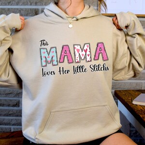 May include: A beige hoodie with the text "This Mama Loves Her Little Stitchs" in black. The word "Mama" is in pink and blue with a heart pattern.