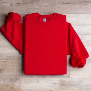 Puede incluir: Una sudadera roja de cuello redondo doblada sobre una superficie de madera. La sudadera es lisa y no tiene ning&uacute;n dise&ntilde;o.