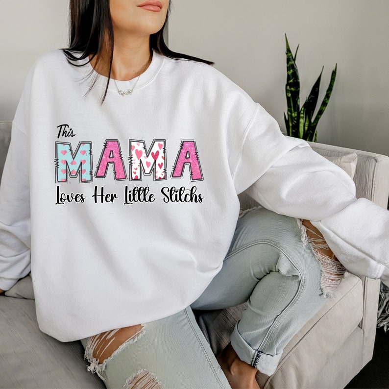 May include: White sweatshirt with the text "This Mama Loves Her Little Stitches" in pink and blue letters with hearts.