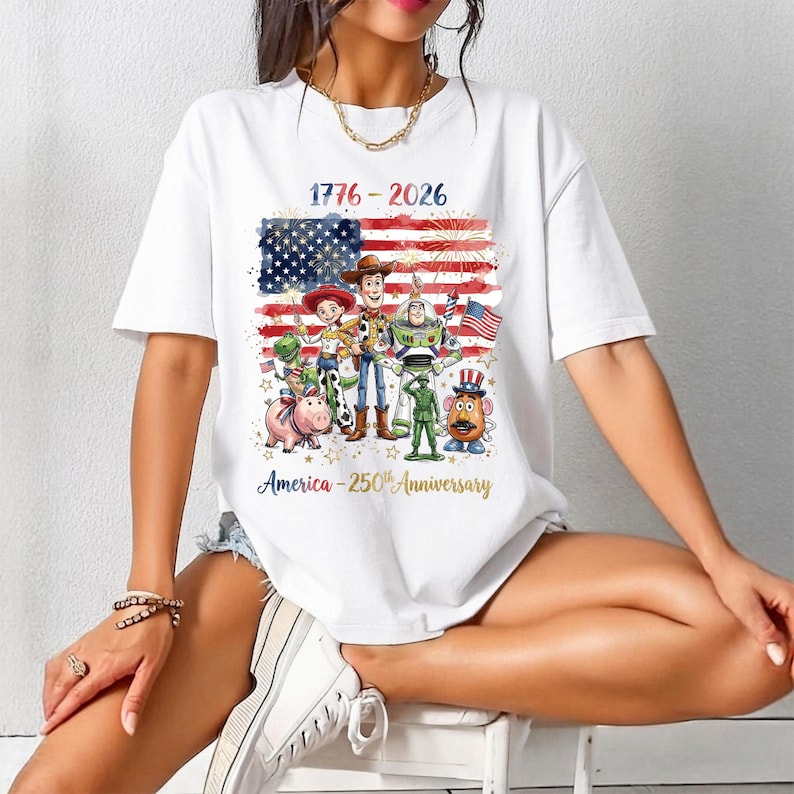 Op de afbeelding: Wit T-shirt met een kleurrijke afbeelding van animatiefiguren en een ontwerp van de Amerikaanse vlag. De tekst op het shirt luidt "1776-2026" en "America - 250th Anniversary" in gouden letters.