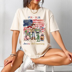 Op de afbeelding: Cr&egrave;mekleurig T-shirt met een afbeelding van de Amerikaanse vlag, vuurwerk en cartoonfiguren. De tekst "1776-2026" en "America - 250th Anniversary" zijn op het shirt gedrukt. Het ontwerp bevat personages uit de Toy Story-franchise.