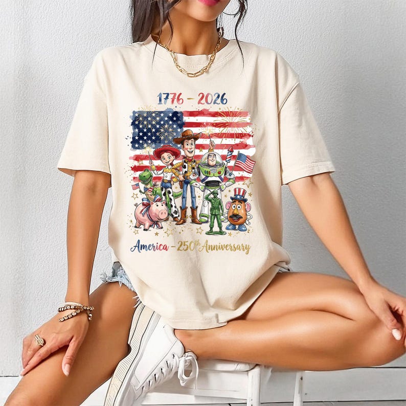 Op de afbeelding: Cr&egrave;mekleurig T-shirt met een afbeelding van de Amerikaanse vlag, vuurwerk en cartoonfiguren. De tekst "1776-2026" en "America - 250th Anniversary" zijn op het shirt gedrukt. Het ontwerp bevat personages uit de Toy Story-franchise.