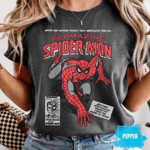 May include: Dark gray t-shirt featuring a vintage comic book cover design of Spider-Man. The graphic includes the text "the AMAZING SPIDER-MAN" in red and white, with additional text and a small image of Peter Parker.