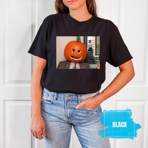 May include: A black t-shirt with a graphic of a pumpkin with a face on it. The pumpkin is on a person's head. The text "BLACK" is in a blue box.