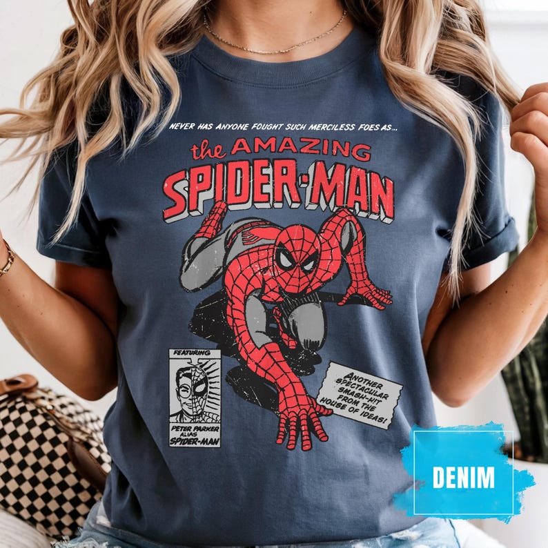 May include: Denim blue t-shirt featuring a vintage comic book graphic of Spider-Man. The design includes the text "the AMAZING SPIDER-MAN" and additional comic book elements. The shirt has a classic, distressed look.