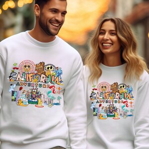 May include: White sweatshirts featuring a colorful graphic design with the text "CALIFORNIA ADVENTURE". The design includes a Ferris wheel, cartoon characters, and other California-themed illustrations. The sweatshirts are worn by two people.