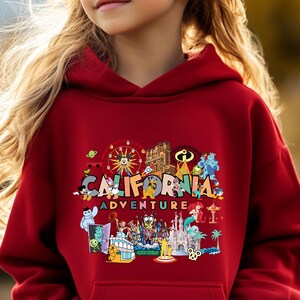 May include: A red hooded sweatshirt featuring the words "CALIFORNIA ADVENTURE" in a colorful design. The design includes cartoon characters, a Ferris wheel, and a castle. The sweatshirt is made of a soft material.