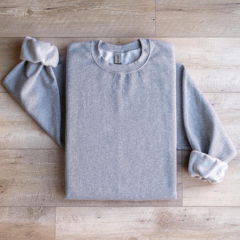 Puede incluir: Una sudadera gris lisa con una etiqueta en blanco. La sudadera est&aacute; doblada y colocada sobre una superficie de madera.