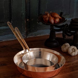 May include: Two copper-bottomed pans with brass handles are nested on a wooden surface. A vintage scale holds shallots, and two garlic bulbs are nearby. The background features a dark green door.