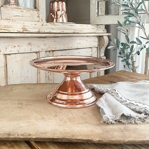 May include: A copper cake stand with a scalloped edge and a pedestal base. The stand is on a wooden surface.