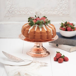 May include: A white cake with a white glaze and fresh strawberries on top, sitting on a copper cake stand. The cake is on a white table with a silver cake server and a white napkin.