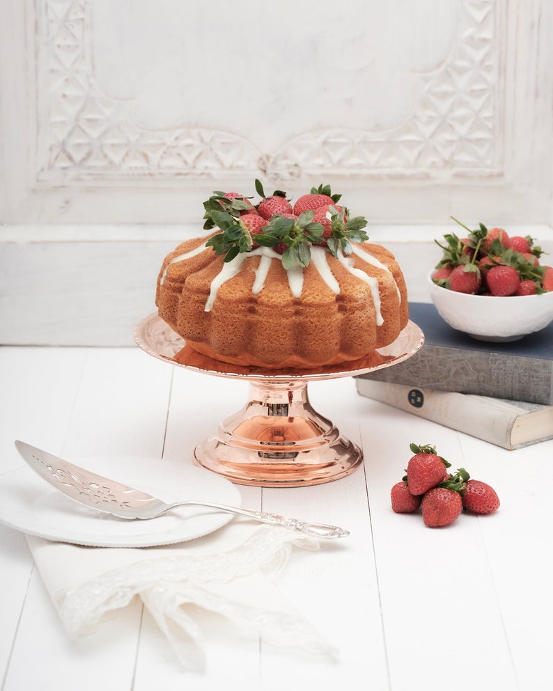 May include: A white cake with a white glaze and fresh strawberries on top, sitting on a copper cake stand. The cake is on a white table with a silver cake server and a white napkin.