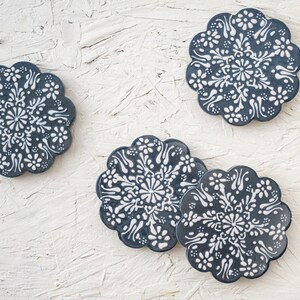 May include: Four navy blue ceramic coasters with a floral design in white. The coasters have a scalloped edge and are arranged on a textured white surface. The floral pattern is symmetrical and detailed.