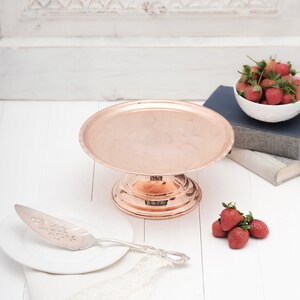 May include: A copper cake stand with a scalloped edge and a decorative base. A silver cake server is on a white plate next to the stand. There are strawberries on a white surface.