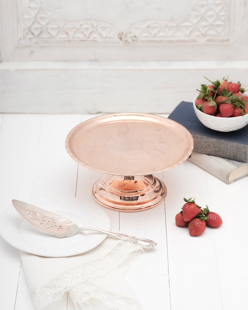 May include: A copper cake stand with a scalloped edge and a decorative base. A silver cake server is on a white plate next to the stand. There are strawberries on a white surface.