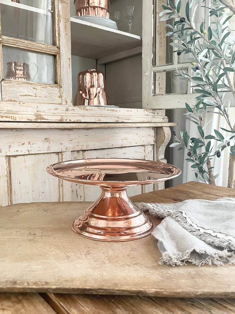 May include: A copper cake stand with a scalloped edge and a pedestal base. The stand is on a wooden surface.