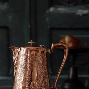 May include: A polished copper coffee pot featuring an intricate embossed botanical pattern of ferns and flowers. The vessel has a curved handle, a tapered spout with decorative detailing, and a small lid with a white knob at the very top center.