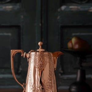 May include: A polished copper coffee pot featuring an ornate embossed floral pattern around the body, a curved handle, a tapered spout, and a small spherical finial on the lid. The metal surface reflects light, highlighting the intricate details.