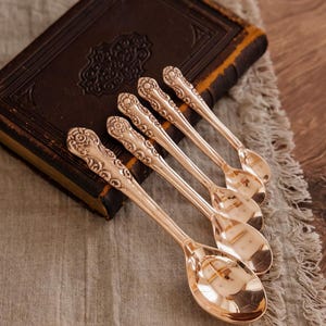 May include: Set of six rose gold teaspoons with ornate handles. The spoons are arranged on a brown leather-bound book with a linen fabric background.