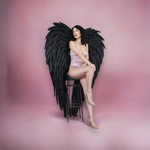May include: A woman sits on a metal stool, wearing a pink bodysuit and beige heels, with large black angel wings behind her. The wings are detailed and textured, set against a pink backdrop.