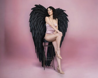 Könnte beinhalten: Eine Frau sitzt auf einem Metallhocker, trägt einen rosa Body und beige Absätze, mit großen schwarzen Engelsflügeln hinter sich. Die Flügel sind detailliert und strukturiert, vor einem rosa Hintergrund.