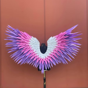 Könnte beinhalten: Ein Paar großer, farbenfroher Flügel mit einem geschichteten Design. Die Flügel weisen einen Farbverlauf auf, darunter Lila, Rosa und Weiß. Die Flügel sind auf einem schwarzen Mannequin-Torso montiert und vor einem einfarbigen orange-braunen Hintergrund ausgestellt.