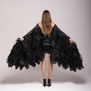 Könnte beinhalten: Eine Person trägt ein schwarzes Paillettenkleid mit einem schwarzen Federumhang. Der Umhang besteht aus vielen dünnen Stoffstreifen, die einen dramatischen Effekt erzeugen.