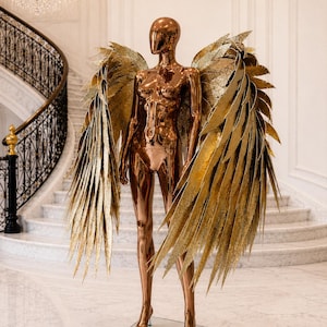 Op de afbeelding: Een bronzen mannequin met grote, goudkleurige vleugels. De vleugels zijn gemaakt van meerdere lagen goudkleurige, puntige veren. De mannequin staat op een glazen voet. De setting is een grote hal met een wenteltrap.