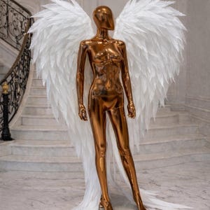 May include: A metallic copper-colored mannequin with large white feathered wings. The mannequin is posed in a standing position on a mirrored surface. The background features a marble staircase and a chandelier.