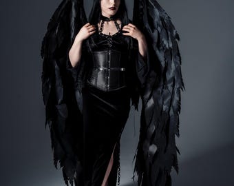 May include: A person in a black gothic outfit with large black feathered wings. The outfit includes a black corset, choker, and long black dress. The person is wearing black heels. The background is a solid gray.