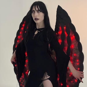 Op de afbeelding: Een persoon draagt een zwarte jurk en grote zwarte vleugels met rode LED-verlichting. De jurk heeft lange mouwen en een split aan de zijkant. De vleugels hebben een geschulpte rand.