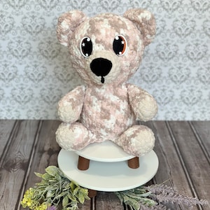 May include: A plush pink and white teddy bear with large black eyes and a black nose sits on a white wooden stool. The bear is sitting on a wooden floor with green and purple flowers.