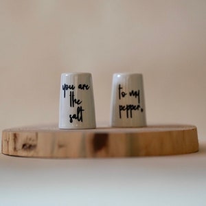 Könnte beinhalten: Weiße Salz- und Pfefferstreuer aus Keramik mit schwarzer Beschriftung. Der Salzstreuer trägt die Aufschrift "You are the salt" und der Pfefferstreuer "To my pepper."