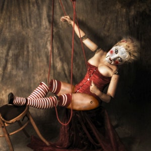 Könnte beinhalten: Eine Frau, die als Marionette verkleidet ist, hängt an roten Schnüren von der Decke. Sie trägt ein rotes Korsett, weiß-rote gestreifte Strümpfe und hat Clown-Make-up im Gesicht. Sie sitzt auf einem Holzstuhl, der mit einem roten Stoff bedeckt ist.