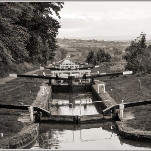 Puede incluir: Una fotografía en blanco y negro de un sistema de esclusas de canal con múltiples compuertas. Las compuertas están hechas de madera y metal y tienen un patrón de rayas blancas y negras. La esclusa está rodeada de árboles y hierba.