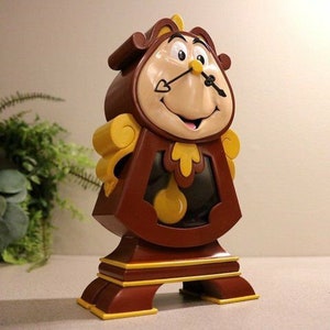 May include: A brown and yellow clock figurine with a smiling face and a large nose. The clock has a pendulum and a face with Roman numerals. The clock is a character from the Disney movie Beauty and the Beast.