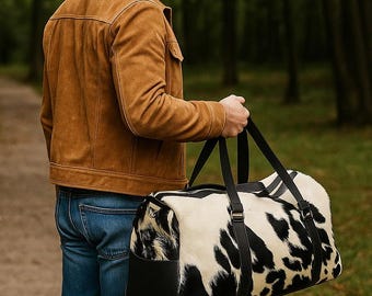 Könnte beinhalten: Eine Person trägt eine Reisetasche mit einem schwarz-weißen Kuhfellmuster. Die Tasche hat schwarze Griffe und Akzente. Die Person trägt eine braune Wildlederjacke und blaue Jeans. Der Hintergrund ist ein Wald.