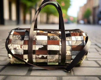 May include: A patchwork duffel bag with a mix of brown, black, and white cowhide squares. The bag has a brown leather shoulder strap and two top handles. The bag is sitting on a brick surface.